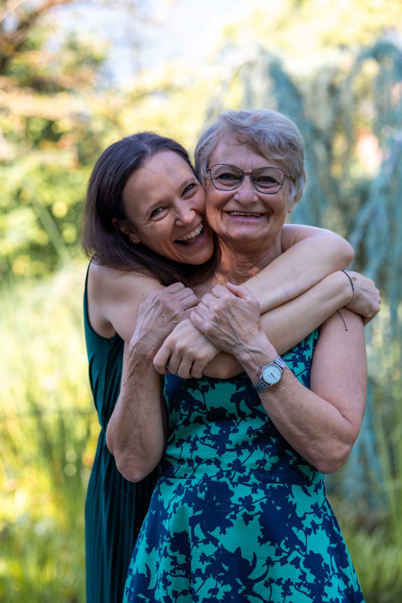 Ältere Mutter wird von erwachsener Tochter umarmt. Beide lachen in die Kamera